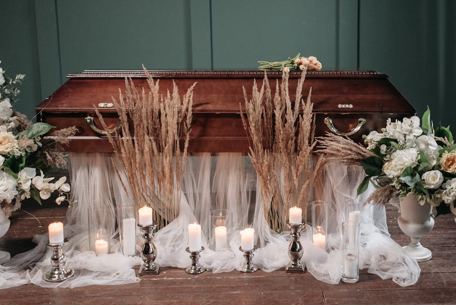 A wooden casket is decorated with floral arrangements including white roses, lilies, and greenery, flanked by tall pampas grass stems. Surrounding the casket are various candles in silver holders and