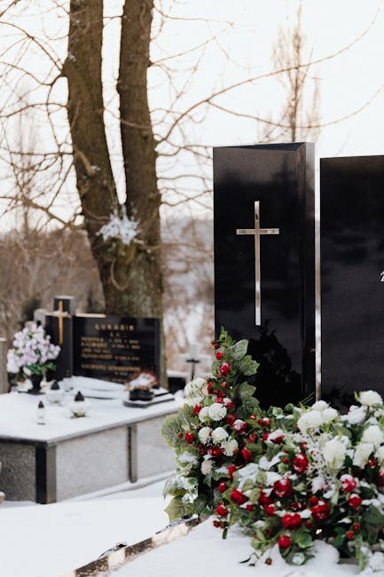 A floral arrangement at a cemetery featuring fresh, vibrant flowers including white roses, red berries, and green foliage, placed in front of a polished black gravestone with a silver cross. The arran