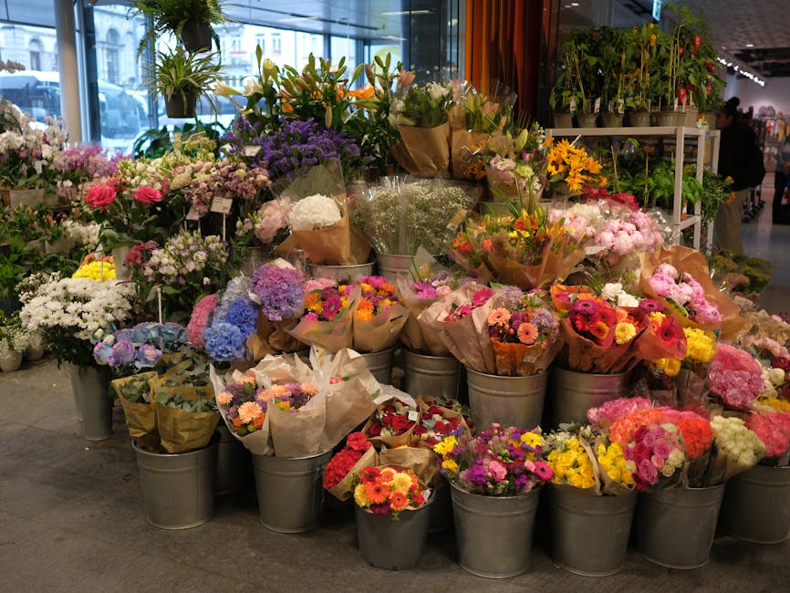 An indoor floral display at a flower shop, featuring a variety of fresh flower bouquets arranged in metal buckets and wrapped in brown paper. The flowers include vibrant pinks, purples, yellows, white
