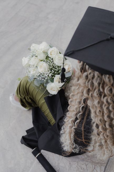 A floral gift arrangement featuring a bouquet of white roses, baby's breath, and green foliage, held by a person dressed in green and black attire, with curly blonde hair and wearing a black cap, plac