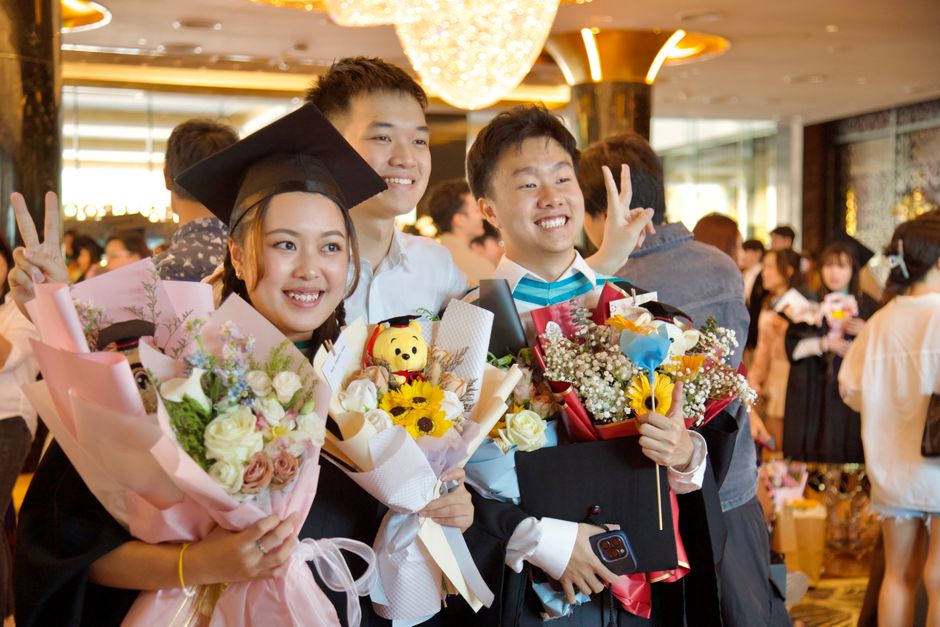 A group of three young adults celebrating at a graduation event indoors, with warm ambient lighting and elegant decor. The woman in the center, wearing a graduation cap, holds a large pink bouquet com