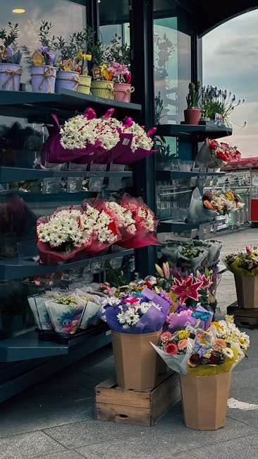 A vibrant display of fresh flower bouquets arranged on black metal shelves and in brown paper wrapping outside a florist shop. The arrangements include a variety of blooms such as white chrysanthemums