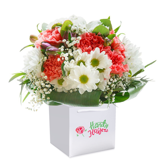 Poised against a spotless white background and cradled within a chic, square white gift bag, this bouquet captures the authentic vibrancy and warmth of New Cross. At its heart, plush coral and deep red carnations bloom, their intricate ruffled textures signifying richness and care. Bright, white daisies emerge amidst the bouquet, their radiant lime-green centers bringing a cheerful sparkle, much like the lively community gatherings found at New Cross's Market Yard. Airy stems of delicate baby's breath weave between the major blooms, infusing the arrangement with softness and visual lift, while a handful of unopened, purple-kissed buds suggest new beginnings and the ever-changing spirit of SE14. Deep green, broad leaves offer a dramatic frame-an homage to the lush canopy in Fordham Park-while also giving structural grace and support to every flower. The bouquet itself rounds out into a generous, harmonious shape, efficient yet abundant, perfect for gifting during significant milestones or simply as an act of everyday kindness within the local area. Dual white ribbon handles trim the bag, hinting at ease of carrying this thoughtful gift on campus or to nearby friends. Luminous lighting ensures every exquisite detail stands out, highlighting the meticulous craftsmanship that New Cross's florists bring to every delivery and occasion.