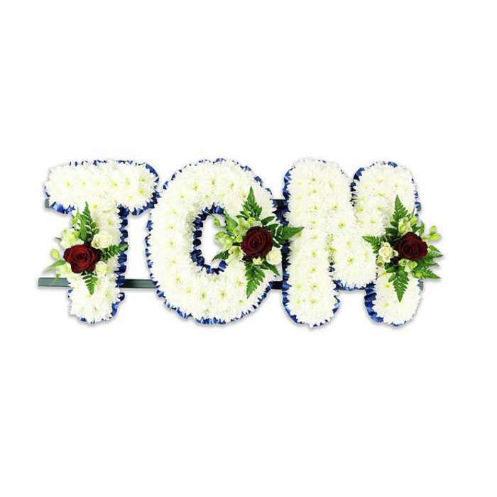 Bold, compassionate and carefully constructed, this funeral tribute spells TOM in large three-dimensional letters entirely covered with tiny white chrysanthemum-like blooms that give each letter a soft, tactile volume. The perimeter of every character is finished with a vivid royal blue ribbon that crisply separates the white field from its surroundings, reinforcing the letterforms with a dignified edge. Inside each letter, a balanced floral spray features one dark red rose as the principal eye-catcher, symbolising deep affection and sorrow; the red is tempered by clusters of pale ivory spray roses and trailing fern fronds that inject a gentle, leafy texture. A thin, dark support bar is visible beneath and behind the letters, indicating the arrangement is designed to stand upright for viewing on an easel or platform. Photographed against a stark white background under soft, diffused light, the composition reads with striking clarity and the layered petals create subtle shadows that lend a soothing three-dimensionality.
Made with the attentive hand of a local floral studio, this tribute would suit memorial gatherings in New Cross - from small chapel services to readings near Goldsmiths or informal wakes by Telegraph Hill Park. It communicates both formality and tenderness: the clean whites and blue border for peaceful dignity, the red roses for personal love, and the green foliage for life remembered. The arrangement feels like a steady, elegant embrace meant to centre a room and comfort those who gather to say goodbye.
