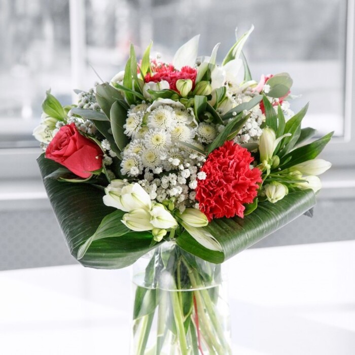 A refined red and white floral arrangement is captured in this photograph, poised in a tall, clear cylindrical glass vase that showcases every detail from stem to petal. The water inside looks freshly poured, with the green stems cut with precision and fanning out neatly, giving the sense that this bouquet has just been arranged by an attentive florist familiar with the pace of life around New Cross Station. Above the rim of the vase, deep red roses stand confidently, their velvety petals gently unfurling, each bloom at that perfect stage between bud and full open flower. Their rich colour brings an immediate feeling of romance and depth, ideal for a dinner at home overlooking the rooftops near Telegraph Hill. At the front, a large, intricately ruffled crimson carnation adds a focal point of texture, its densely layered petals catching the light and adding a sense of movement. Surrounding these central red tones are soft white and cream accents: Alstroemeria with slim, slightly curved petals etched with delicate speckles, and several smooth, tightly closed white lily buds that suggest beauty yet to come, extending the life and interest of the bouquet day by day. Fine sprays of gypsophila - baby's breath - are threaded throughout, their tiny white flowers resembling a dusting of snow, cushioning the bolder blooms and adding an ethereal lightness. The bouquet is held together visually by lush greenery; broad, glossy dark green leaves are curled and wrapped around the base like an elegant collar, while slimmer eucalyptus-like sprigs and pointed leaves introduce varied shapes and cooler green tones. The arrangement rests on a clean white surface in front of a softly blurred, bright backdrop, perhaps a sunlit New Cross living room or café window. The overall effect is one of polished warmth and gentle celebration, perfect for anniversaries, Mother's Day, or as a gracious gift to bring comfort and joy to someone nearby. Crafted with the care you'd expect from Flower Delivery New Cross, it feels both contemporary and timeless.