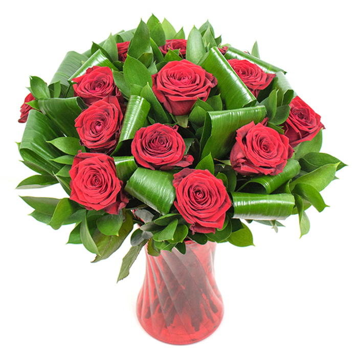 A striking floral study in crimson and green: roughly a dozen or more fully opened deep red roses form a tightly clustered, spherical bouquet whose velvety petals spiral inward in perfect, opulent curls. The intense red of each bloom ranges from true crimson at the petal faces to richer, almost wine-toned shadows where petals overlap, and the even, soft lighting reveals delicate petal veins and that plush, tactile surface. Interspersed through and beneath the rose heads is a wealth of dark, glossy foliage-smaller, dense leaves that produce a verdant backdrop and broader, shiny leaves that have been folded and rolled into elegant, tubular shapes. Those rolled leaf forms-crafted with an eye for modern floral architecture-cradle the roses and introduce sweeping arcs and sculptural rhythm into the arrangement. The bouquet is anchored in a tall, translucent ruby-red glass vase with a subtle ribbed or twisted finish; its vertical texture refracts light, elongates the silhouette, and intensifies the red theme while giving a firm, refined base to the display. Presented against a neutral white surface, every contour and hue reads crisply: the greens read darker, the reds glow richly, and the composition reads as both classic and contemporary. The overall effect is romantic and sophisticated, the soft rose perfume implied by the close-packed blooms making it ideal for anniversaries, Valentine's Day, or an intimate celebration. As a local touch, I might imagine this on a side table near Brockley Rise or delivered to a Georgian terrace along New Cross Road for someone who appreciates traditional blooms with a modern twist.