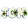 This custom funeral tribute is composed of three large floral letters that spell TOM, designed as a central display piece for services in and around New Cross. The letters are built in solid block shapes, with the visible surfaces completely covered in tightly packed white chrysanthemums, creating a smooth, cushiony texture that looks soft and serene. Their pure white petals form a quiet, unbroken background that speaks of peace and remembrance. Around the perimeter of each letter, a royal blue satin ribbon is skillfully pleated, giving a crisp, tailored edge that sharply defines the name and adds a touch of formality, ideal for a church or crematorium setting. At chosen points on every letter-the top of the 'T', the side of the 'O', and part of the 'M'-small sprays of accent blooms are worked into the chrysanthemum base. These include velvety deep red roses, smaller creamy spray roses, and slim, elegant white flowers reminiscent of freesia, arranged together and framed by glossy greenery and delicate fern fronds that arc gently over the white cushion. The tribute is supported by a slim dark frame running beneath the letters, hidden enough to keep the focus on the flowers while keeping the piece secure whether placed by the coffin or at the front of a service hall near local streets such as Queens Road. The overall combination of white, blue, red, and green gives this floral name arrangement a dignified yet warmly personal character, offering a heartfelt way for family and friends to remember and honour Tom within the close-knit New Cross community.