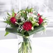 A luxuriant red and white bouquet stands proudly in a clear cylindrical glass vase, its clean lines echoing the modern homes and studios around New Cross. Through the transparent glass, you can see the freshly cut green stems fanning out in the water, each one neatly trimmed and carefully placed, suggesting the quiet skill of a local florist who knows how to balance structure and softness. Above the waterline, velvety deep red roses rise as the main feature, their petals unfurling in tight spirals that speak of classic romance and heartfelt passion, ideal for an anniversary dinner on a New Cross Road balcony or a surprise gift after a walk in Fordham Park. At the front of the arrangement, a large, densely ruffled crimson carnation catches the eye, its layers of petals adding rich texture and a touch of drama. Around these bolder blooms, clusters of white and cream blossoms bring contrast and calm. White Alstroemeria, with their slim, speckled petals, mingle with creamy buds of lilies that are still closed, suggesting beauty on the verge of opening, like an early morning in nearby Telegraph Hill Park. Sprays of delicate white gypsophila float among the larger flowers, their tiny stars softening the overall look and creating a gentle, cloud-like fullness. Curved, glossy dark green leaves wrap elegantly around the base, forming a collar that cradles the bouquet, while narrower eucalyptus-like foliage and pointed greens add layers of shape and tone. The arrangement is photographed on a pristine white surface with a softly blurred, bright background, likely a sunlit room. The even light highlights the freshness of every petal and leaf, giving the bouquet a polished yet welcoming mood, perfect for celebrations, birthdays, or simply brightening a New Cross workspace with warmth and affection.