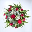 From a high, top-down view, this generously full bouquet appears as a vibrant circle of colour and texture set against a pristine white background, as if freshly placed on a florist's workbench in New Cross. At its centre, velvety deep red roses dominate the eye, some still tightly furled in bud, others just beginning to open to reveal layered crimson petals that suggest romance and heartfelt emotion. Nestled closely around them are ruffled carnations in rich fuchsia and ruby red tones, their plush, frilled petals creating a lively, almost festive energy that echoes the bustle of nearby New Cross Road on a bright morning. Threaded between these warm hues are clusters of small, daisy-like white spray chrysanthemums, each neat bloom adding a clean, luminous contrast that keeps the arrangement feeling fresh and airy. Sprinkled delicately throughout is a cloud of tiny white gypsophila, or baby's breath, its countless miniature florets hovering like a soft mist, filling any gaps and lending the bouquet a dreamy softness. The flowers are framed by a generous collar of greenery: strong, pointed dark green leaves, likely from lilies or alstroemeria, radiate outward to give structure, while rounded, silvery-green eucalyptus leaves curl and overlap, providing a contemporary touch and hinting at a cool, herbal fragrance. The overall composition feels robust yet refined, a carefully balanced hand-tied design that would sit beautifully on a kitchen table overlooking Fordham Park or brighten a flat near Goldsmiths, University of London. This bouquet's vivid reds, crisp whites, and lush greens make it ideal for anniversaries, Valentine's Day, birthdays, or simply sending an uplifting message of love and recovery to someone special in SE14.