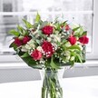 A lush bouquet of rich red roses, matching red carnations, and crisp white alstroemeria stands proudly in a clear glass vase on a bright white tabletop, bathed in gentle natural light from a large window. The roses range from tight, dignified buds to fully open blooms with velvety petals that curl softly at the edges, while the carnations bring a ruffled texture that makes the arrangement feel full and generous. White alstroemeria, some still in bud and others just opening to reveal delicate trumpet-shaped petals with pale green tips, weave between the deeper reds, creating a graceful contrast. Tiny sprigs of white gypsophila, or baby's breath, float like a fine mist through the bouquet, softening the stronger colours and lending a romantic, almost cloud-like lightness. Around the base and threaded throughout, broad, glossy green leaves and finer, lighter foliage form a structured collar, giving the display a polished silhouette and a sense of movement, as though a breeze from nearby Telegraph Hill Park had passed through. Through the clear vase, the neatly trimmed stems can be seen standing in clean water, reinforcing the impression of freshness and care in every detail. The background beyond the window is softly blurred and nearly monochrome, perhaps echoing the muted greys of New Cross Road on a calm morning, so that all attention rests on the vivid reds, whites, and greens at the centre. The overall mood is one of cheerful appreciation and heartfelt celebration, like flowers delivered to a New Cross flat to mark an anniversary, a birthday, or simply to brighten a loved one's day with a confident burst of colour and fragrance.