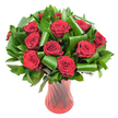 A lush, rounded bouquet of more than a dozen deep crimson roses sits like a jewel in this image, each bloom opened fully to reveal velvety, wine-dark petals that curl inward in tight, luxurious spirals. The rich red surface of every petal catches soft, even studio light, revealing subtle gradients from ruby to burgundy and the fine, satiny texture that invites the fingertips. Surrounding the rose heads is a generous halo of dark green foliage: glossy, flat leaves that form a dense verdant backdrop and broader, emerald leaves artfully rolled and folded into architectural loops and spirals. Those sculpted green shapes-reminiscent of Aspidistra folds and Ruscus collars-give the composition height and modern polish, their shiny faces and crisp edges creating contrast against the roses' plush softness. The stems are gathered and presented in a tall, slender ruby-red glass vase whose translucent surface is subtly ribbed or twisted, throwing vertical lines of light that echo the stems and deepen the overall red tone. Set against a clean white background, the arrangement is isolated to showcase colour and form: the reds appear more saturated, the greens darker and more luminous, and every petal and curvature reads with clarity. As a florist I imagine the faint, classic scent of roses rising from the cluster-a gentle, heady perfume that suits anniversaries, Valentine's gestures, or an elegant centrepiece at a candlelit dinner. This bouquet carries a sense of refined romance and quiet luxury, the kind of floral statement that would look at home on a mantel on New Cross Road or as a thoughtful delivery for someone living near Telegraph Hill Park.