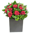 A modern, sculptural bouquet of rich, velvety red roses arranged as a tight cluster at the centre, each bloom opening in soft spirals that catch the light like silk. Broad, glossy green leaves are carefully folded and curled around the roses, forming rhythmic arcs and layered planes of emerald that add movement and visual depth; up close the foliage has a firm, almost waxy texture that contrasts with the petals' plush softness. The composition sits in a low, square black box vase with a matte finish, its geometric silhouette grounding the arrangement and lending a contemporary, gallery-like clarity to the piece. Bright, steady studio lighting highlights the luminous edges of each leaf and the velvet sheen on the crimson petals, suggesting the fresh clarity of morning light on New Cross Road or the crisp interior lighting of a Goldsmiths studio. There is a subtle scent suggestion: the classic sweetness of rose softened by a green, leafy top note-imagine the smell of clipped garden leaves mingling with heady floral warmth. This bouquet reads as both confident and intimate, suited to an anniversary dinner on a quiet terrace near Telegraph Hill Park or as an elegant gift to say thank you after a visit to Deptford Market Yard. The craftsmanship is deliberate and restrained: each curl of foliage and each rose placement feels intentional, a small study in balance and devotion that makes an immediate, tactile impression even before it is smelled. A piece like this communicates romance and refinement without fuss, bringing a quietly dramatic focal point to contemporary homes and urban gatherings in and around New Cross.